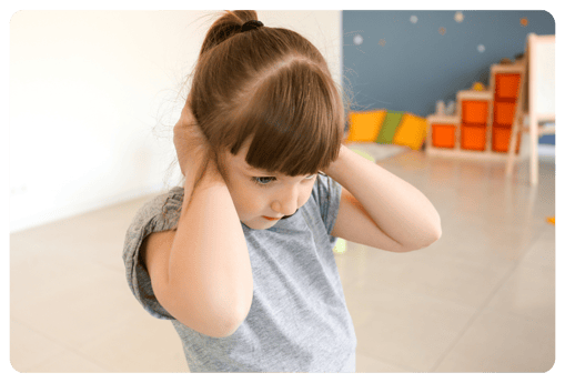 An expressionless six-year old girl wearing a pony tail and a gray t-shirt holding her ears to block sound. 