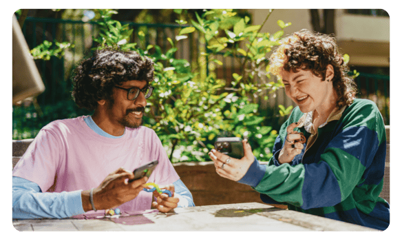 A teenage girl wearing a blue and green striped rugby shirt laughing and showing her phone to a teenage boy wearing glasses. Both are holding colorful puzzles.