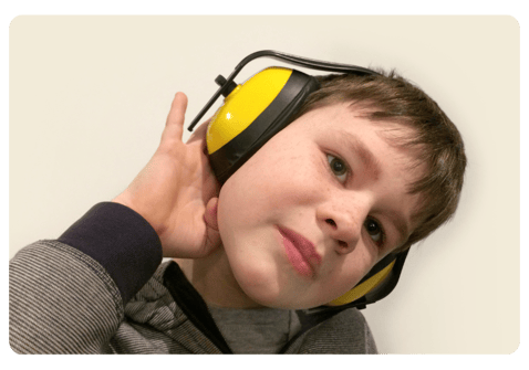 7-year old boy wearing a gray and black stripped hoodie and bright yellow noise-cancelling headphones turning his head and holding his right ear.