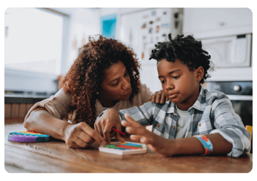 Eight-year old African American boy sitting at a table doing a colorful puzzle with the assistance of his mother, who has her hand on his shoulder and is talking to him.